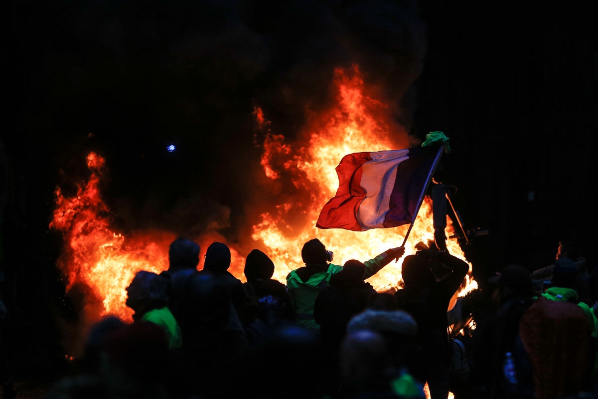 A French flag floats as fire is burning during a protest of Yellow vests (Gilets jaunes) against rising oil prices and living costs, on December 1, 2018 in Paris. - Speaking at the Paris police's command centre, French Prime Minister said 36,000 people we