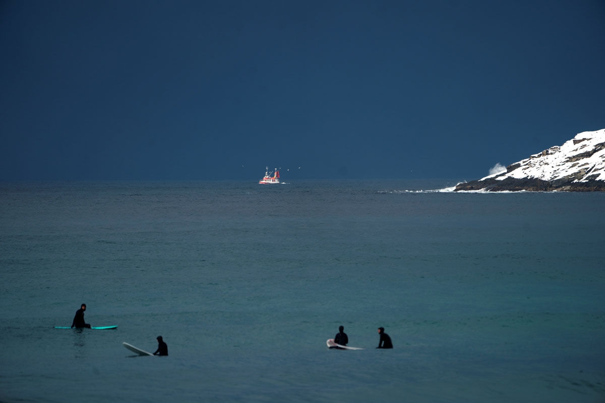 Surfers wait for waves as a fishing boat sails by, on March 11, 2018 in Unstad, northern Norway, Lofoten islands, within the Arctic Circle as air temperature drops minus 13°C and water temperature above 4°C. .