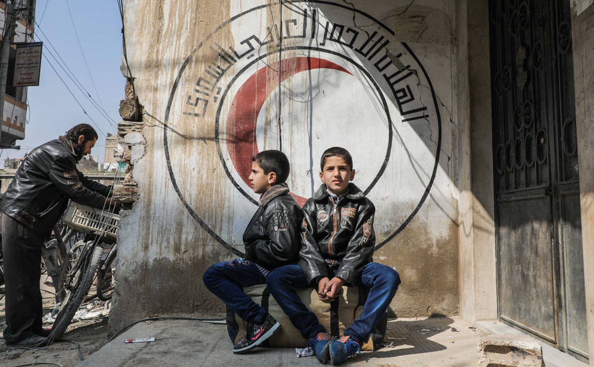 Syrian children sit on a luggage bag before a mural on a wall bearing the logo of the Syrian Red Crescent at the organisation's branch in Douma in the rebel-held enclave of Eastern Ghouta on the eastern outskirts of the capital Damascus on March 13, 2018,